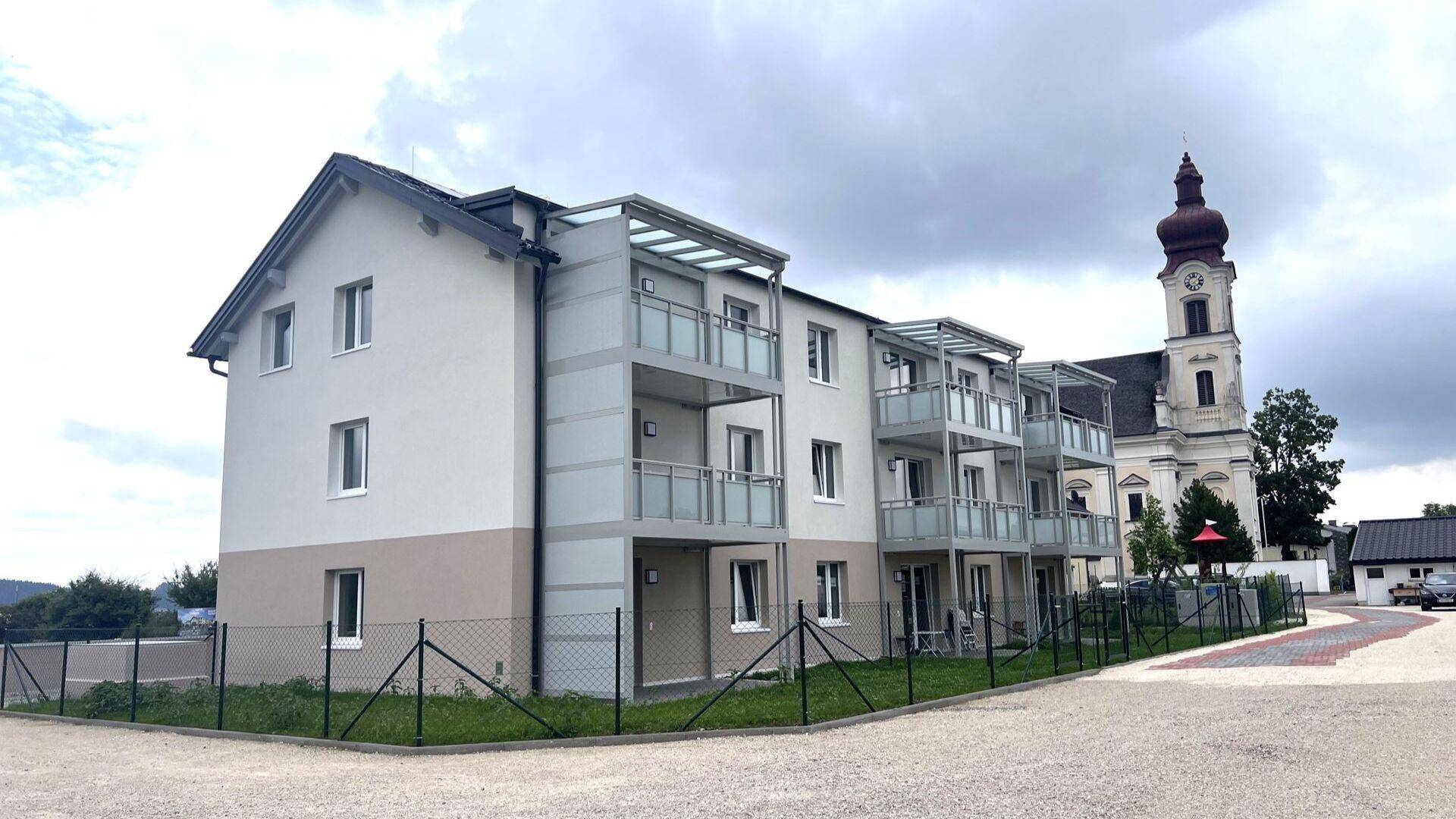 Neubau-Wohnanlage mit Balkonen neben einer Kirche unter bewölktem Himmel.