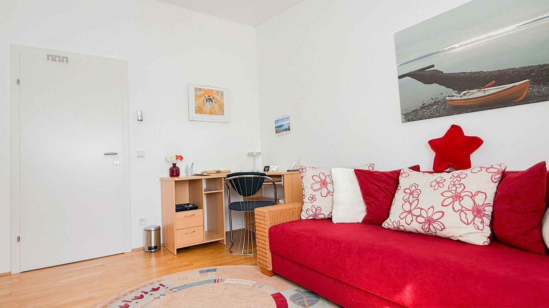 Freundliches Zimmer mit rotem Sofa, Schreibtisch und Holzboden, ideal als Gäste- oder Arbeitszimmer.