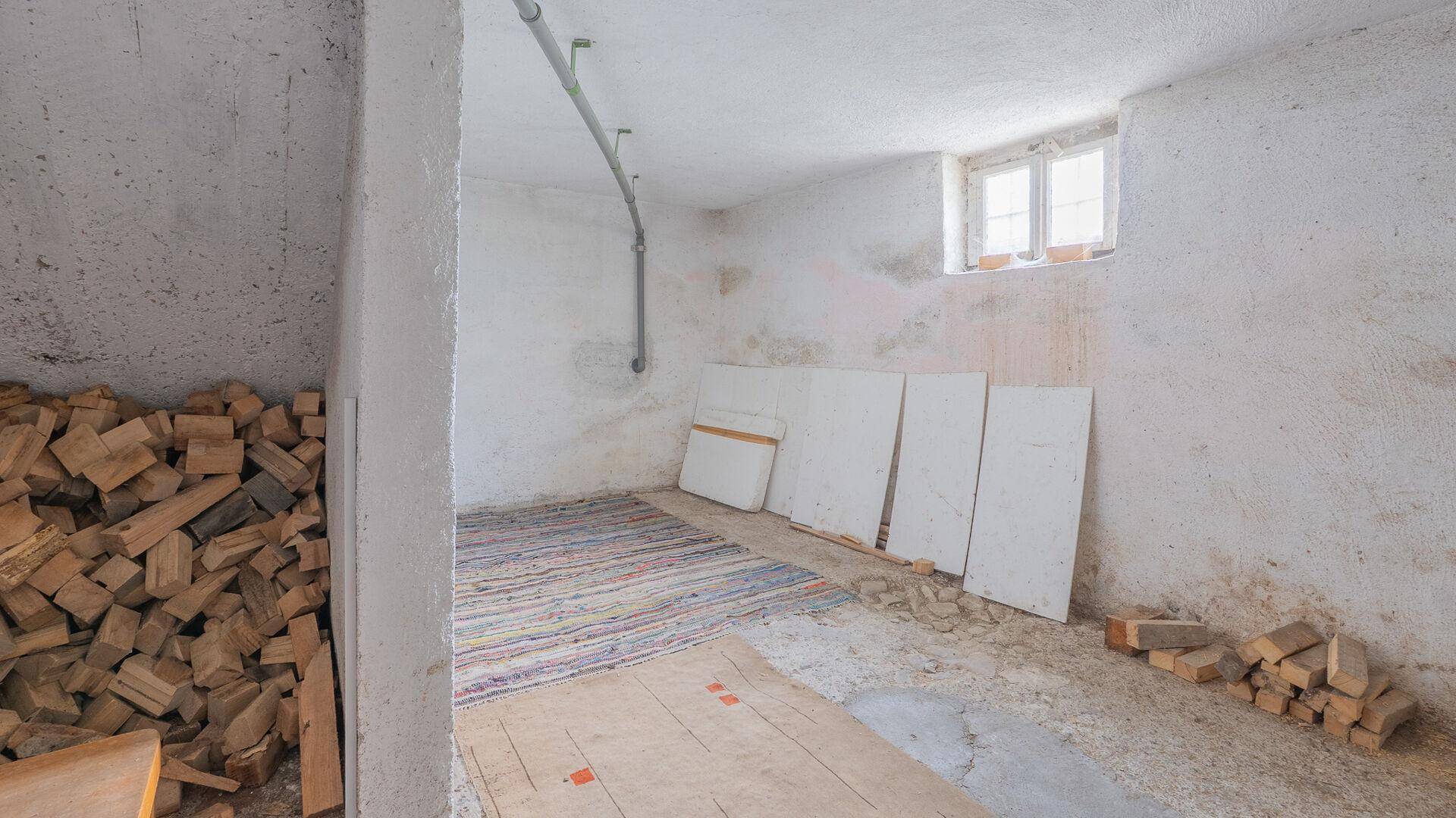 Unausgebauter Kellerraum mit Holzstapel und kleinem Fenster, ideal als Lager.