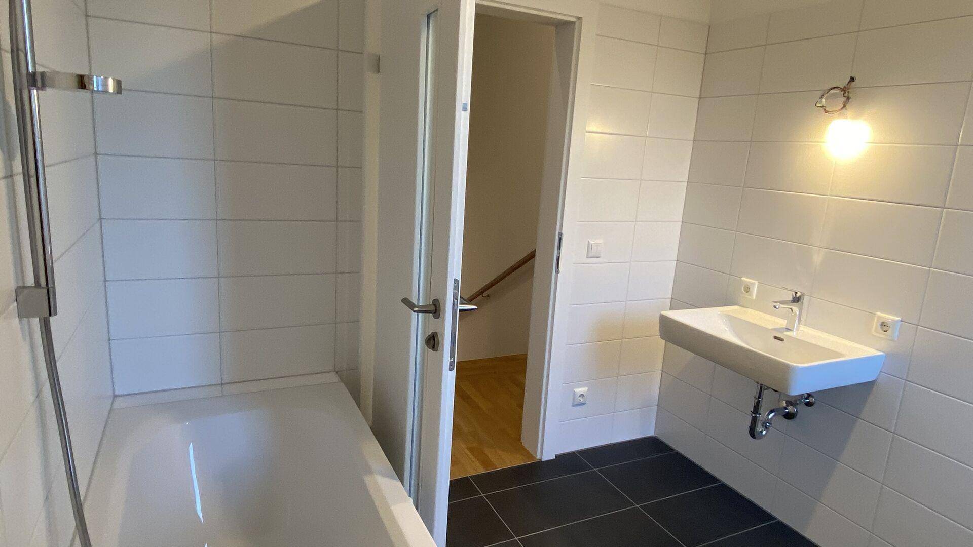 Helles Badezimmer mit Badewanne, Duschfunktion und Waschbecken, Zugang zum Treppenhaus.