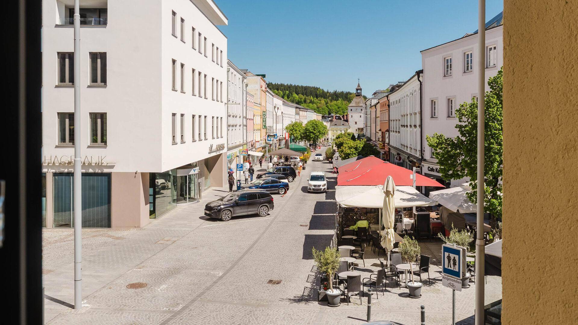 Blick aus einem Fenster auf eine belebte Einkaufsstraße mit historischen Gebäuden und einem Uhrturm.