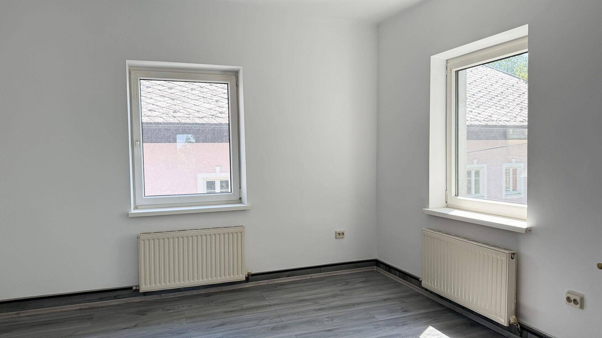 Heller Raum mit weiß gestrichenen Wänden, grauem Laminatboden und zwei Fenstern mit Blick auf Nachbargebäude.