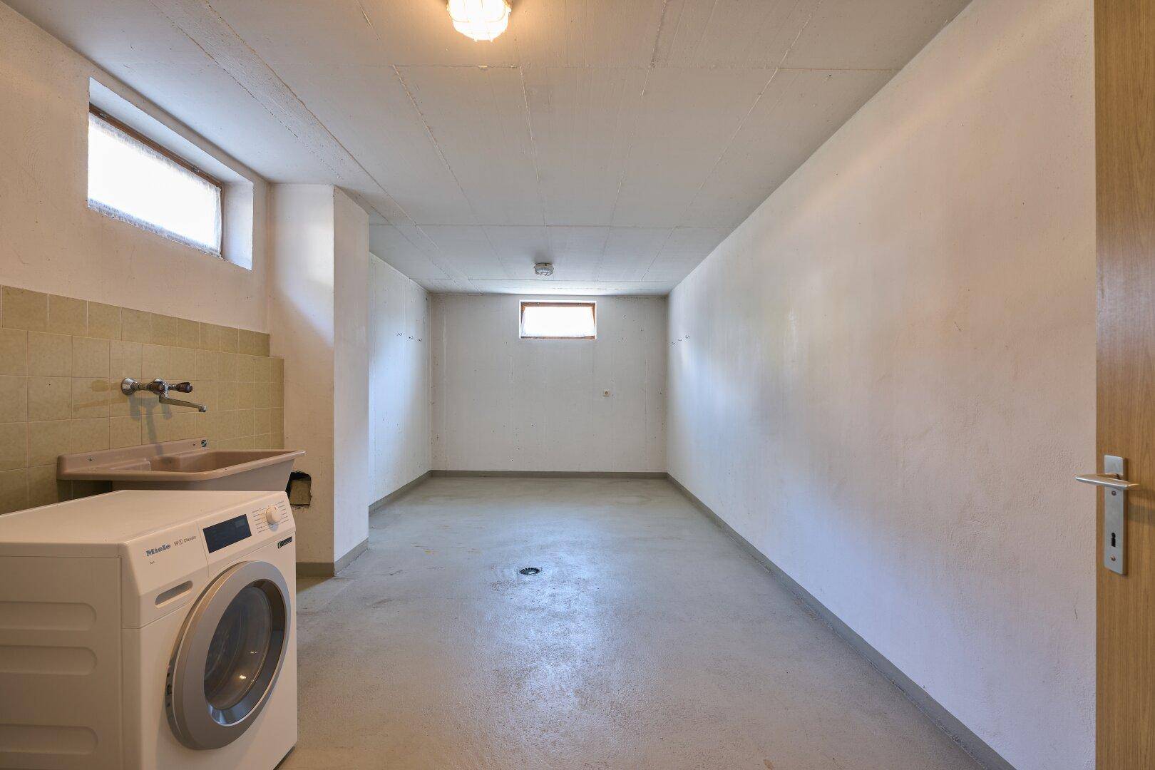 Waschküche im Keller mit Waschmaschine, Spülbecken und kleinem Fenster.