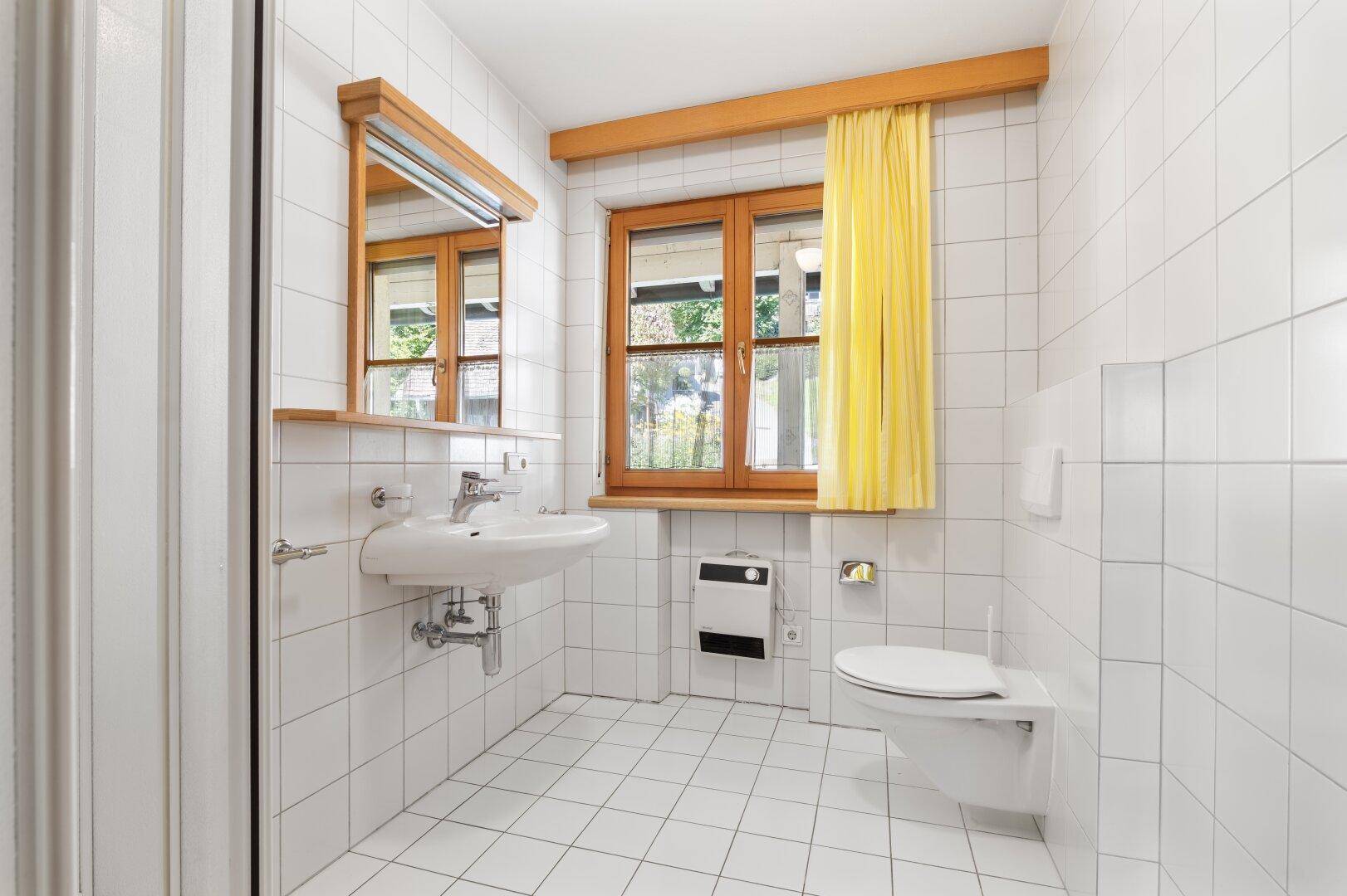 Badezimmer mit weißen Fliesen, Waschbecken, Toilette und einem Fenster für Tageslicht.