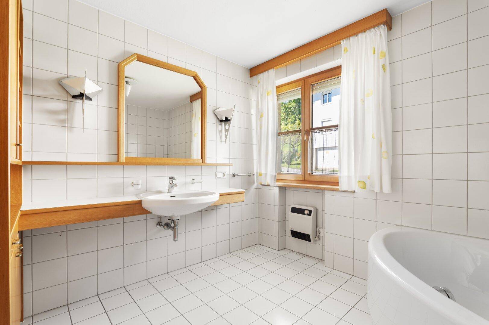 Helles Badezimmer mit weißen Fliesen, Waschbecken, Spiegel und Fenster.