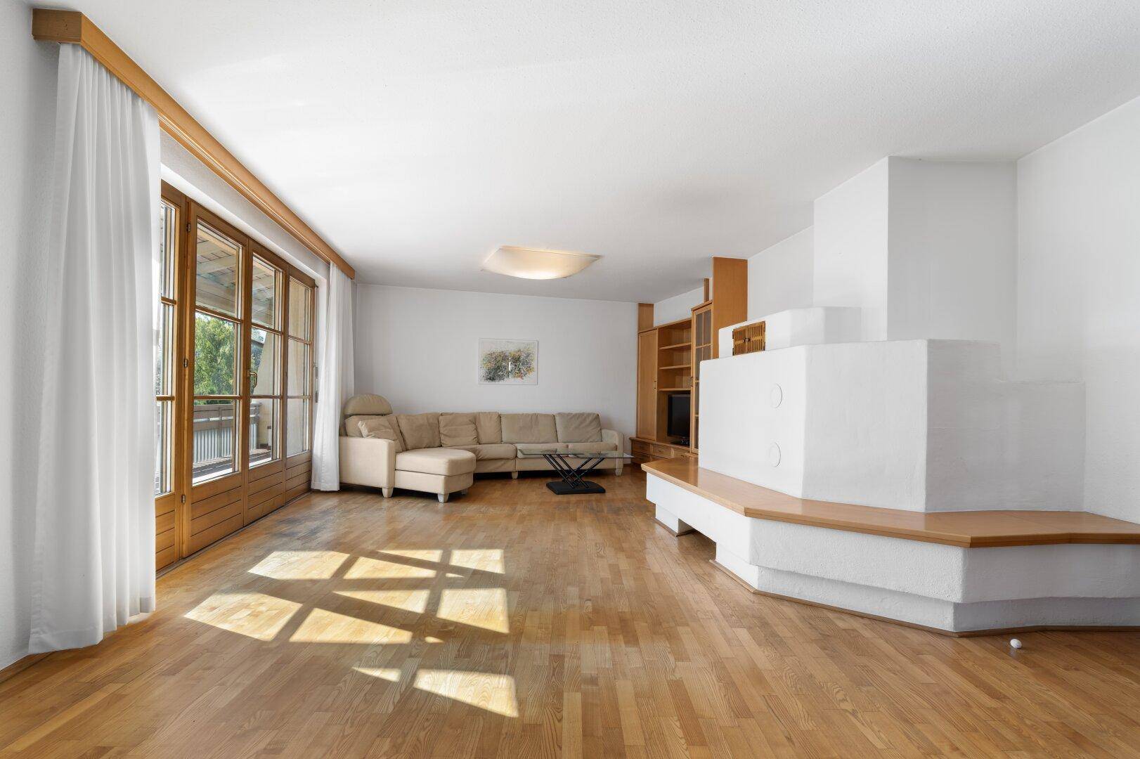 Geräumiges Wohnzimmer mit Holzboden, großem Kachelofen und Zugang zum Balkon.