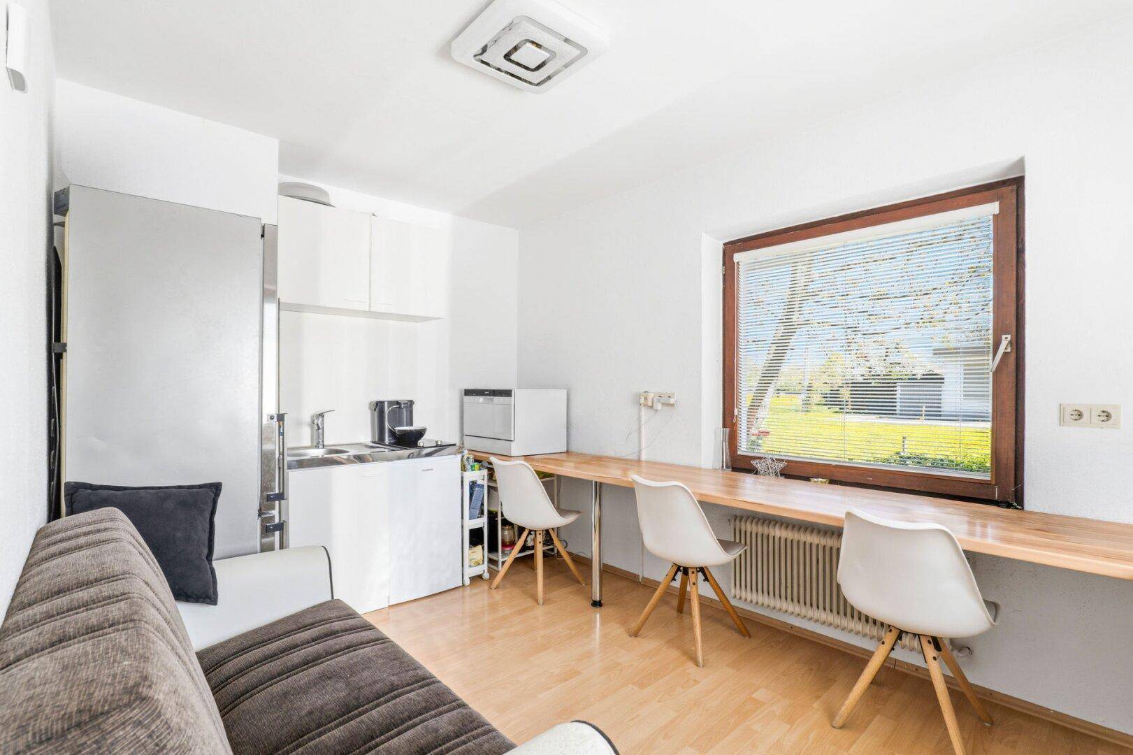 Gemütlicher Wohnbereich mit integrierter Küchenzeile, Esstisch und Sofa, hell durch großes Fenster.