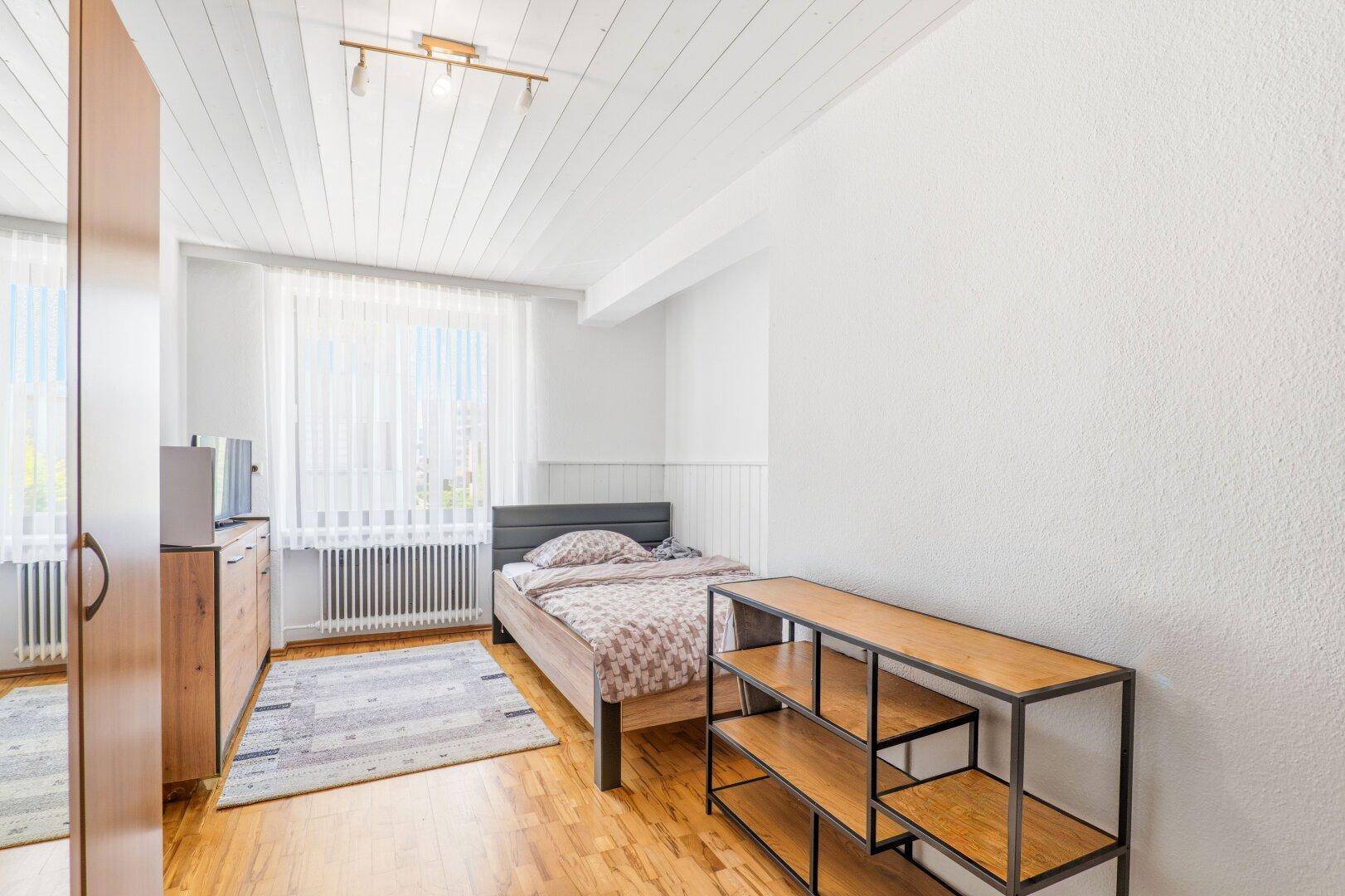 Helles Schlafzimmer mit Holzboden, weißer Decke und einem Einzelbett, gut beleuchtet durch ein Fenster.