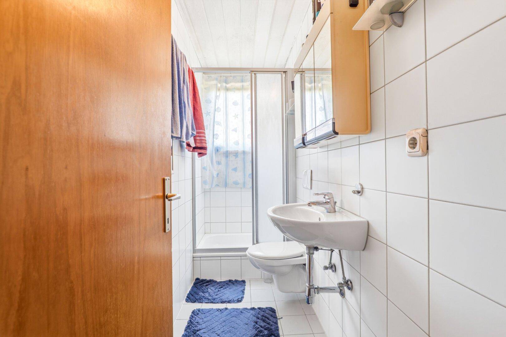 Kleines Badezimmer mit Duschkabine, WC und Waschbecken, komplett gefliest in Weiß.