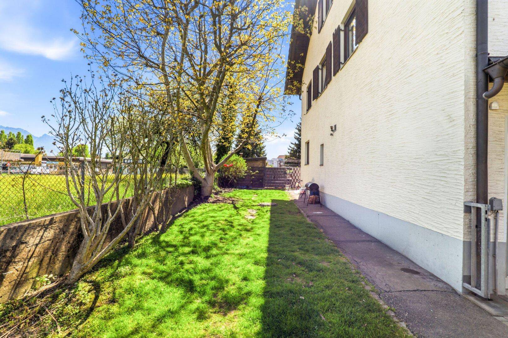 Seitlicher Gartenbereich mit Rasenfläche, Bäumen und einem Zaun, der das Grundstück begrenzt.