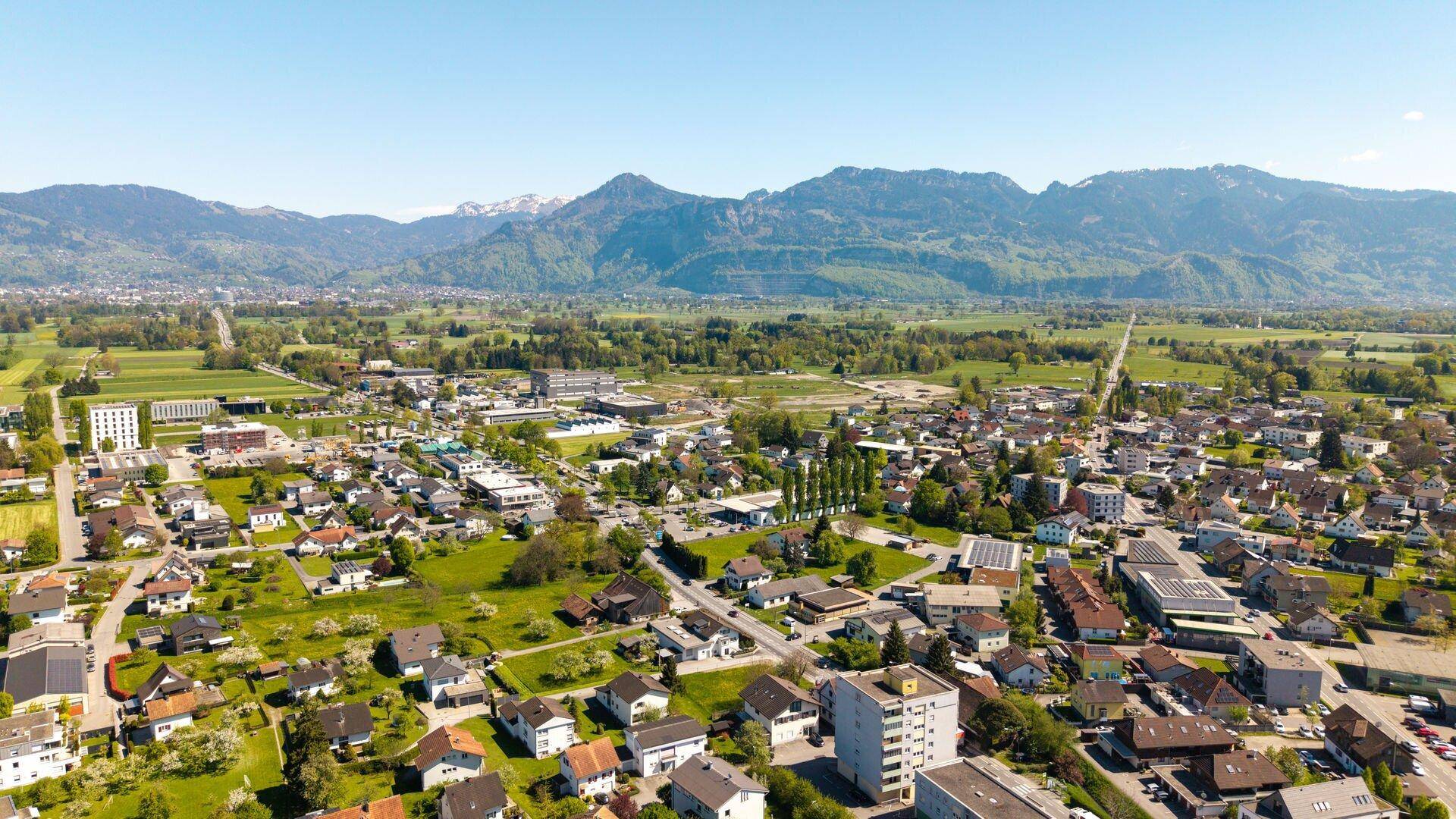Weitläufige Luftaufnahme einer Stadt mit Wohngebieten, Grünflächen und majestätischen Bergen im Hintergrund.