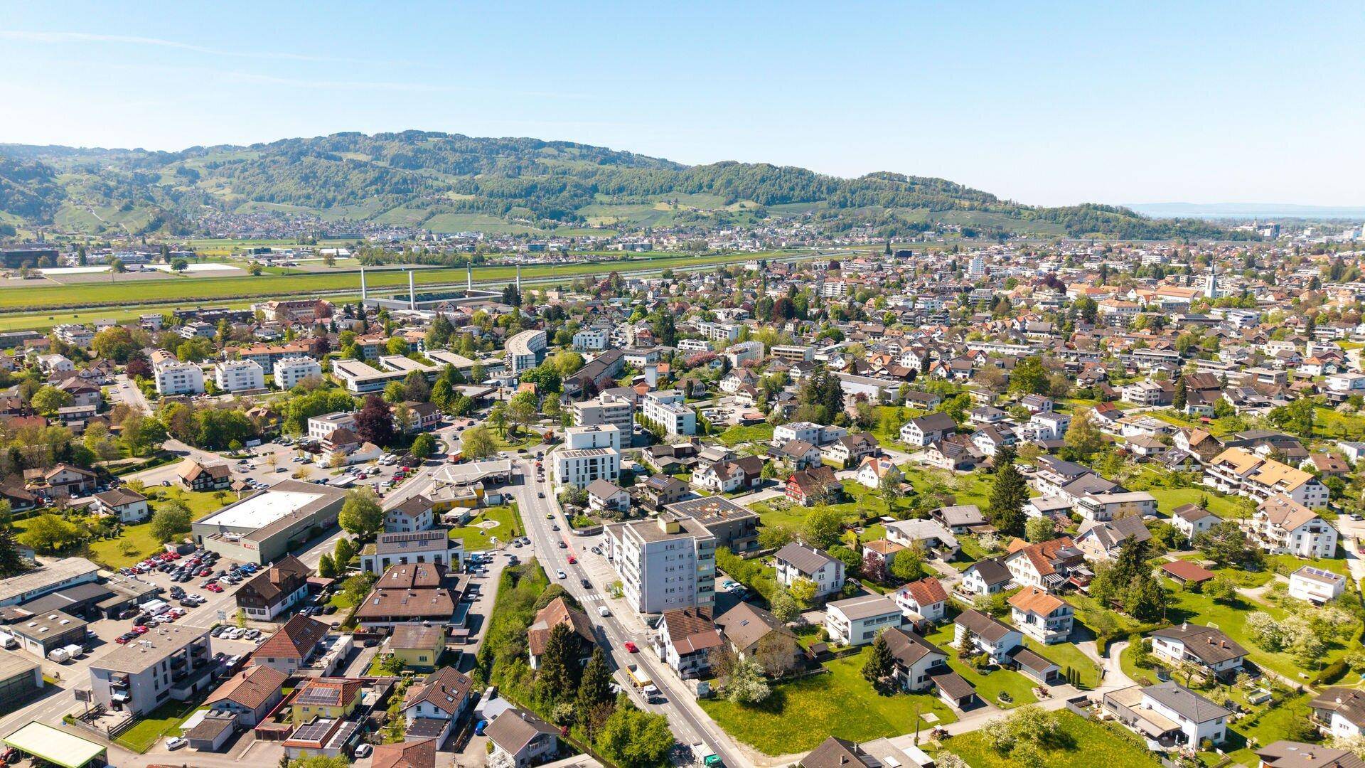 Ausgedehnte Luftaufnahme einer Stadt mit Wohngebäuden, Straßen und bewaldeten Hügeln in der Ferne.