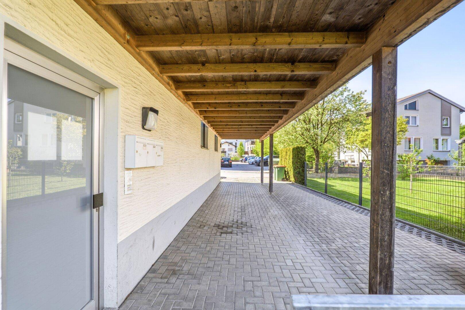 Überdachter Eingangsbereich oder Carport mit Holzbalken und gepflastertem Boden neben dem Gebäude.