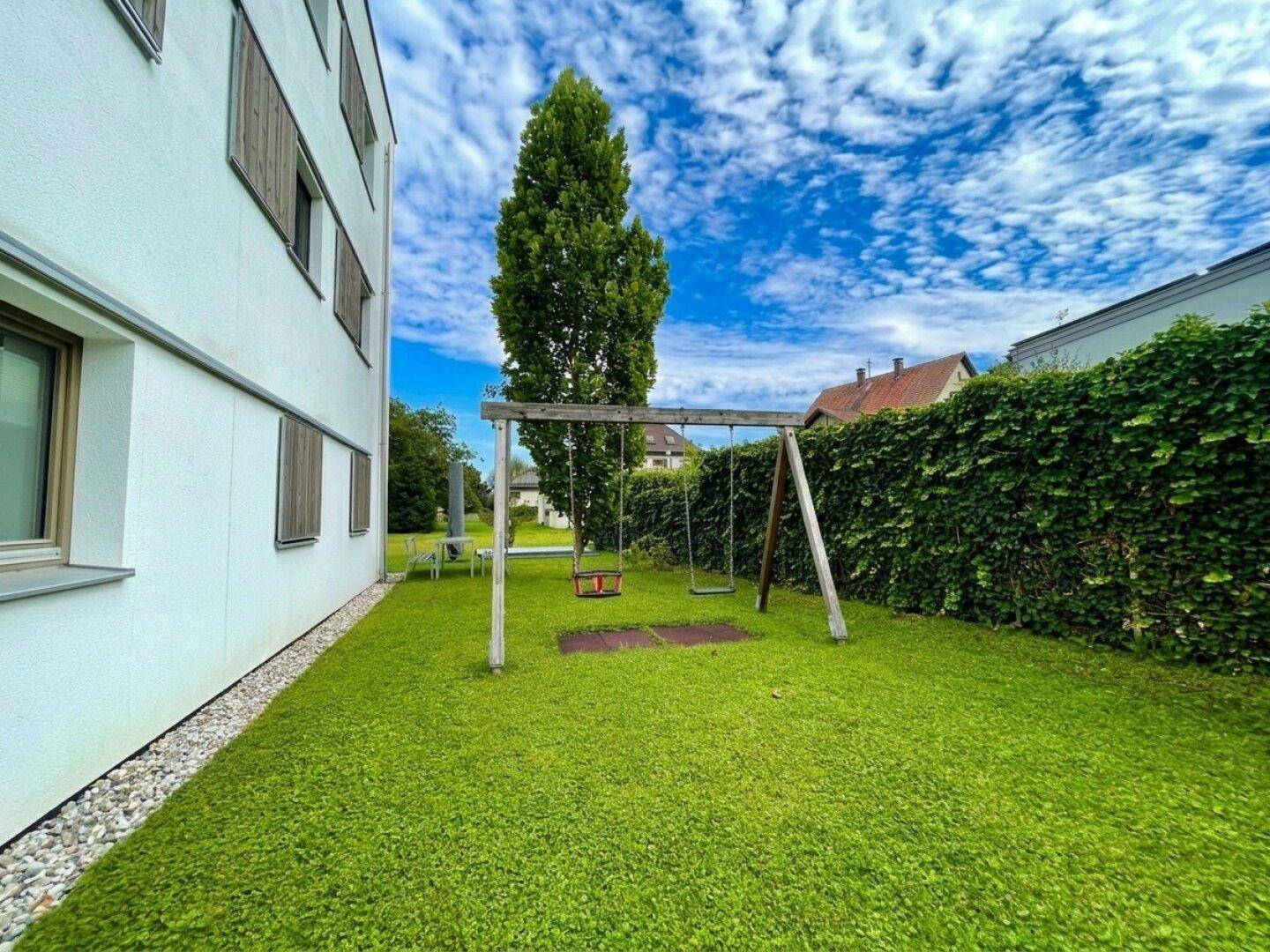 Gemeinschaftsgarten mit Rasenfläche, Schaukel und Hecke neben dem Gebäude.