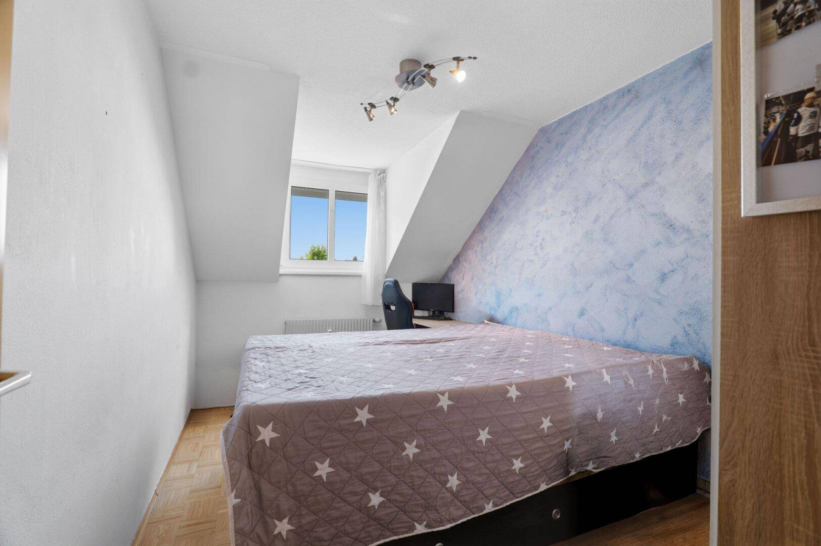 Gemütliches Schlafzimmer mit Dachschräge, blau akzentuierter Wand und Parkettboden.