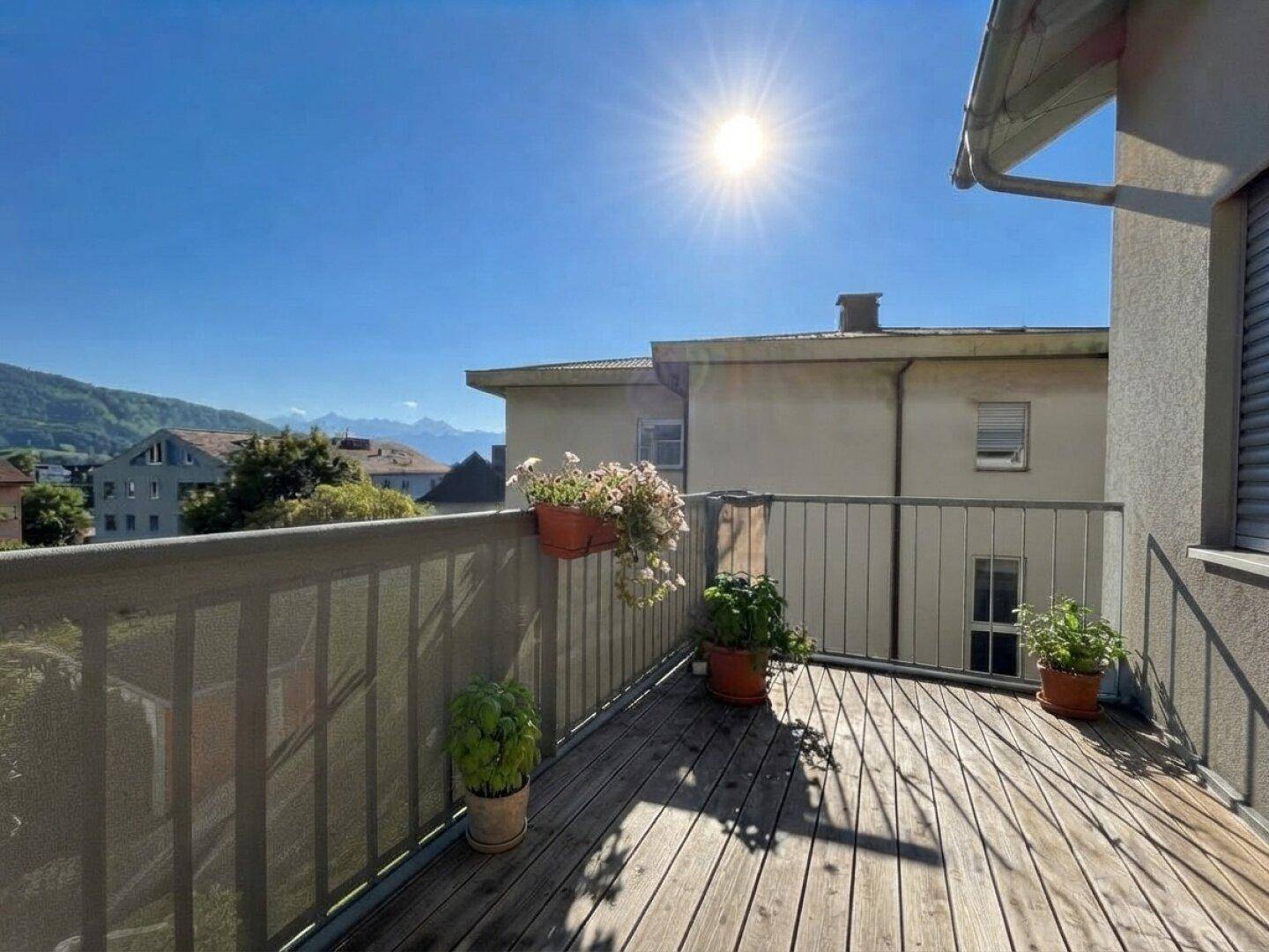 Sonniger Balkon mit Holzboden, Pflanzen und weitem Blick auf die Berge.