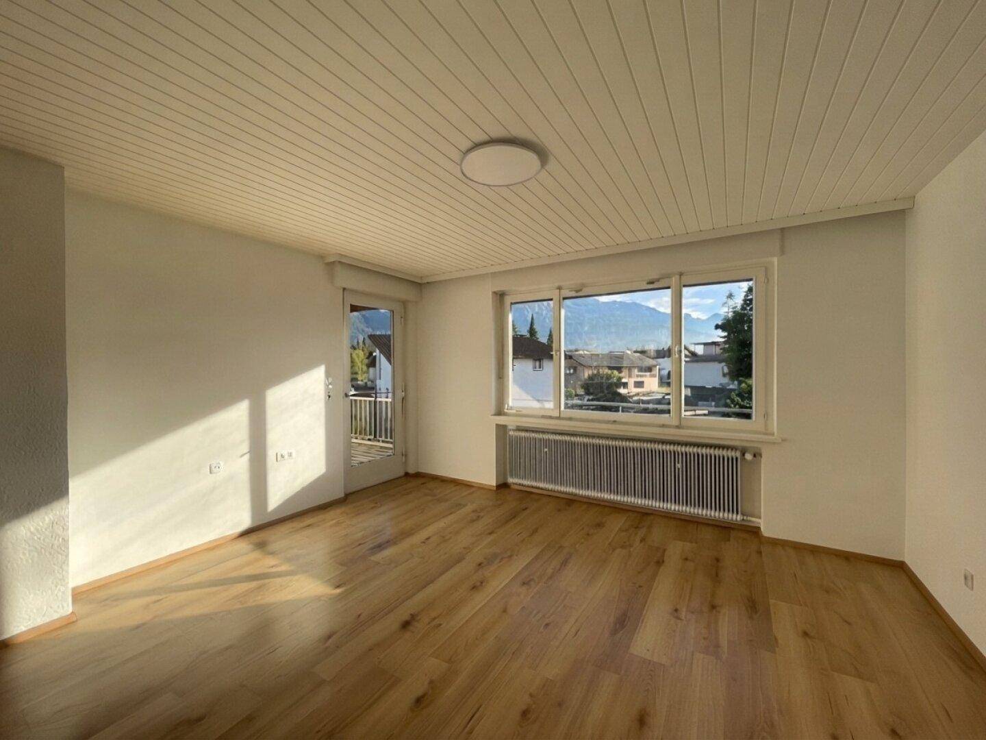 Helles Wohnzimmer mit Laminatboden, großem Fenster und Zugang zum Balkon mit Bergblick.
