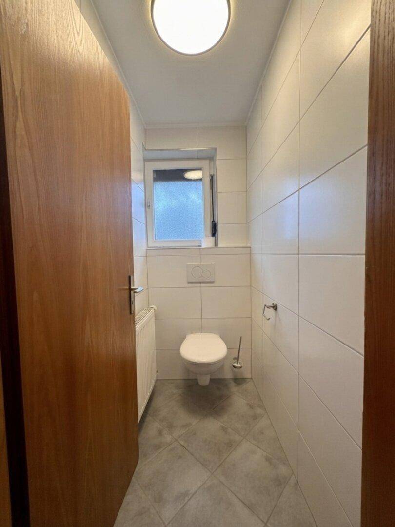 Separates WC mit wandhängender Toilette, weißen Fliesen und kleinem Fenster.