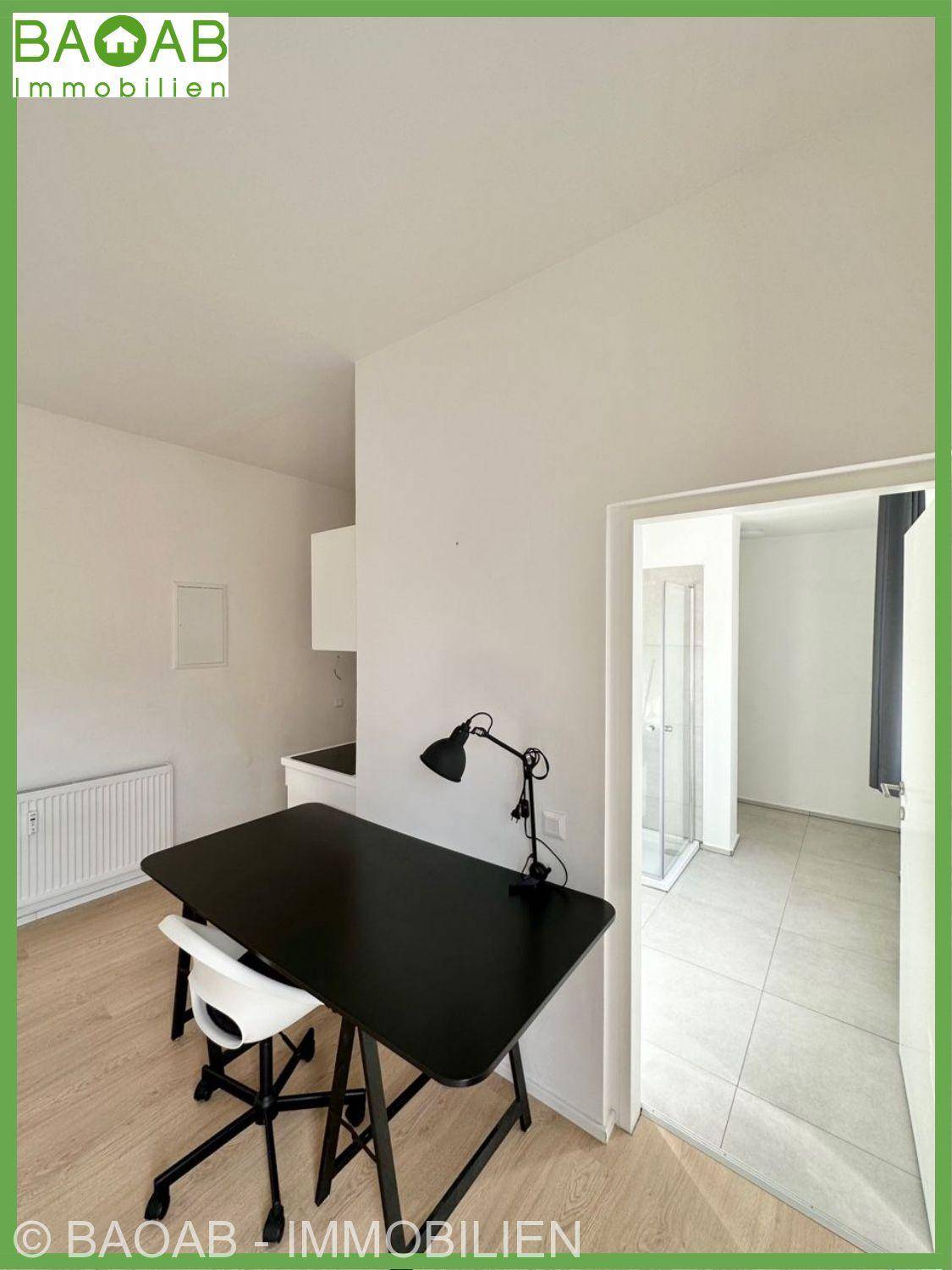 Heller Wohnbereich mit Schreibtisch und Zugang zu einem Badezimmer mit Duschkabine.