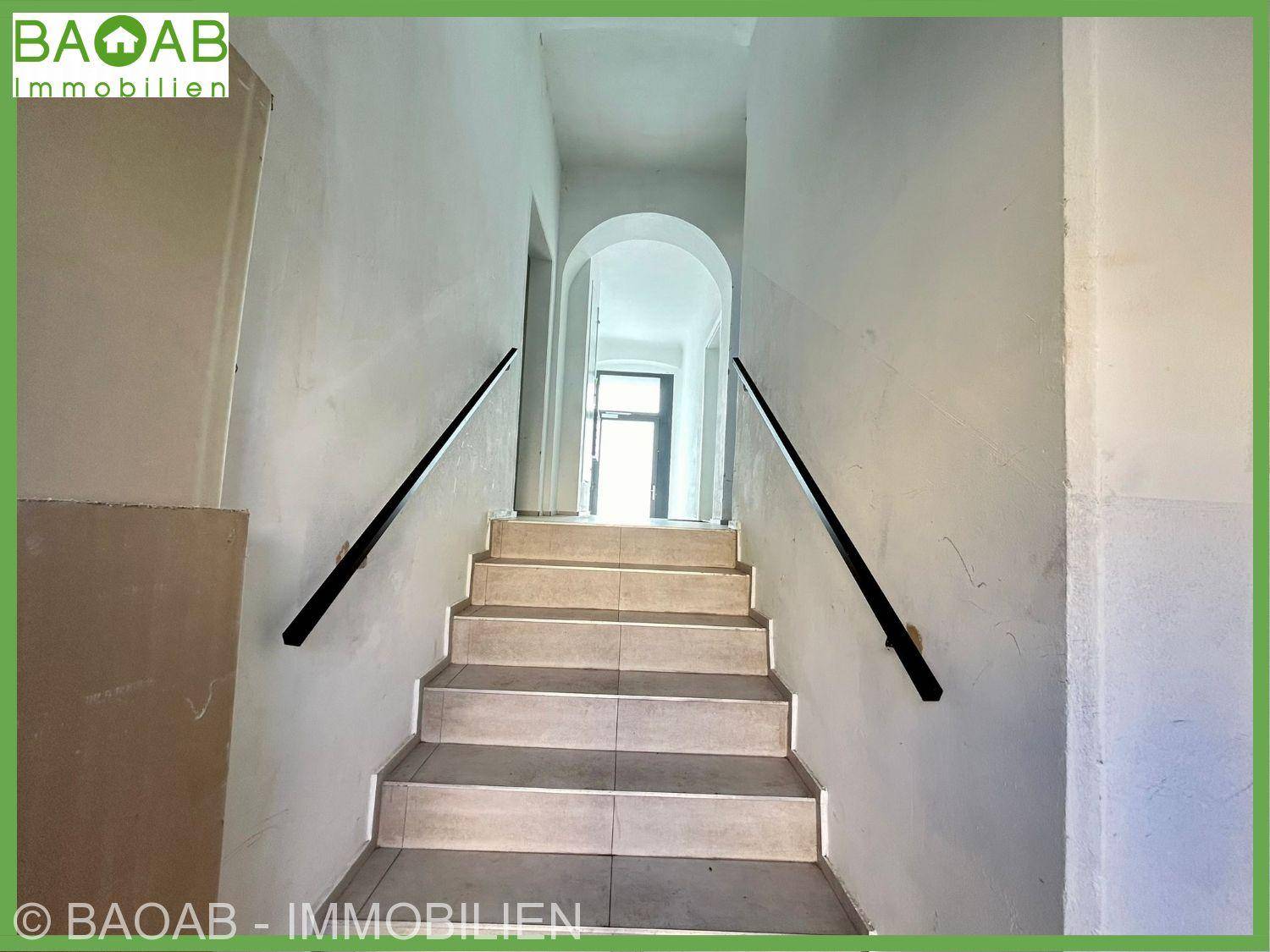 Treppenhaus mit gefliesten Stufen und schwarzen Handläufen, führt zu einem hellen Flur mit Bogen.