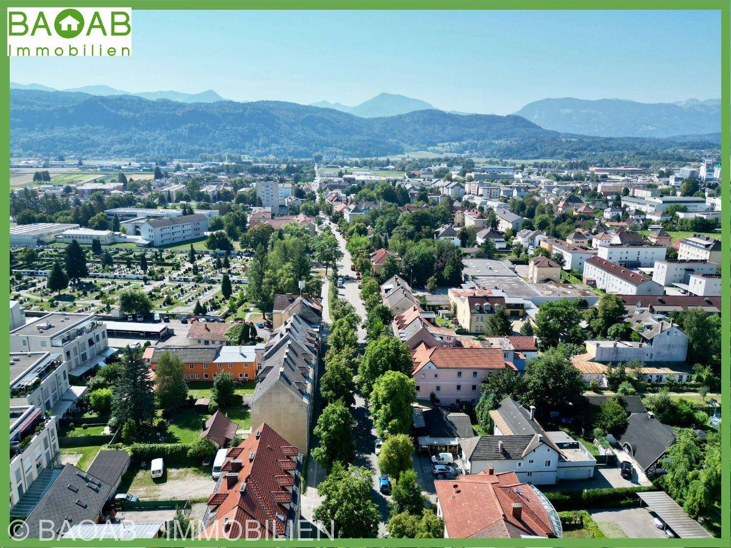 Weitläufige Luftaufnahme einer Stadt mit Wohngebäuden, viel Grün und einer beeindruckenden Bergkulisse.