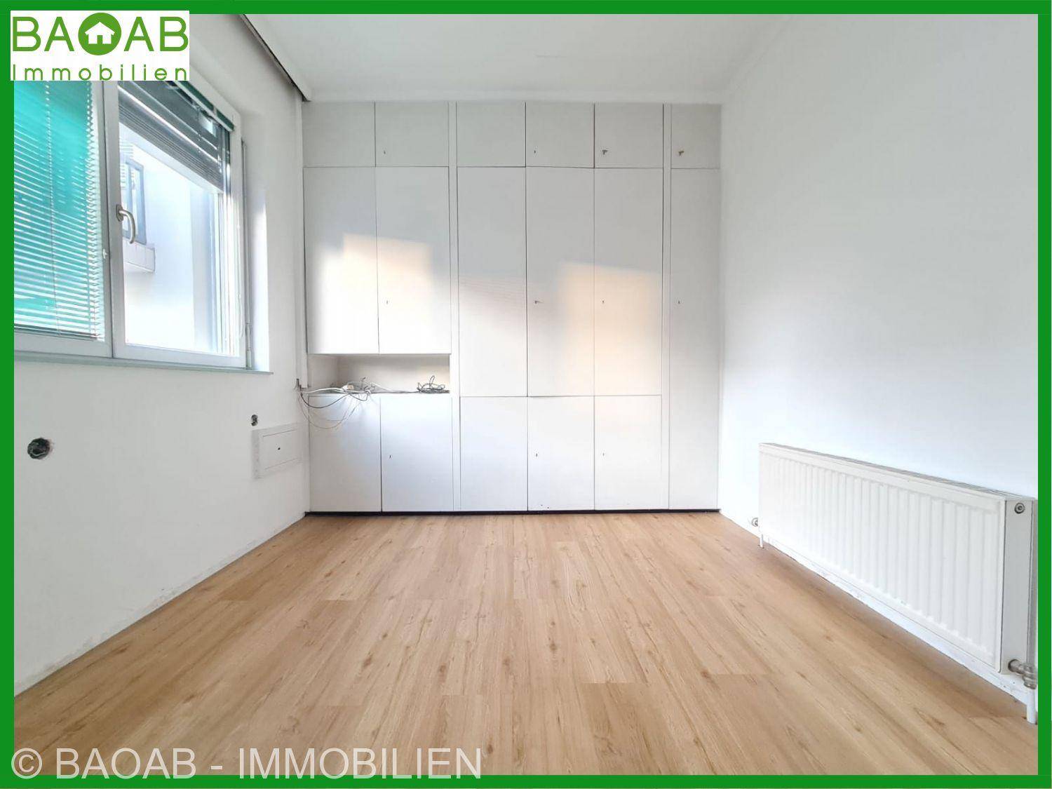 Leerer Raum mit integrierten weißen Schränken, Holzboden und Heizkörper unter dem Fenster.