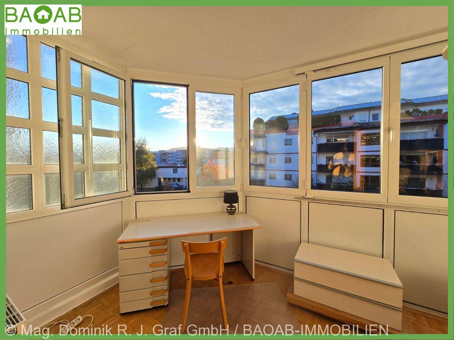 Heller Arbeitsbereich mit Schreibtisch und Stauraum, umgeben von großen Fenstern mit Stadtblick.