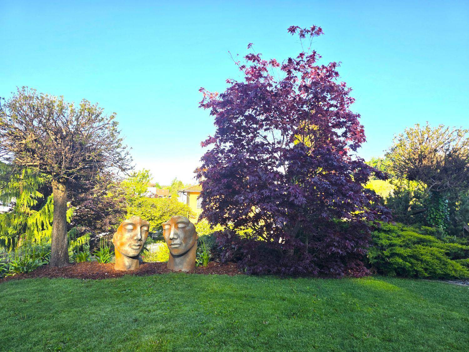 Gepflegter Gartenbereich mit Rasenfläche, Bäumen und dekorativen Skulpturen unter blauem Himmel.