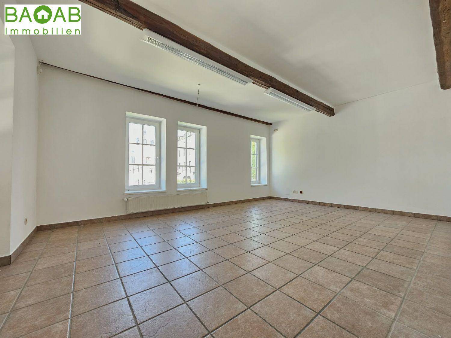 Leerer Büroraum mit Fliesenboden, weißen Wänden, drei Fenstern und einem sichtbaren Holzdeckenbalken.