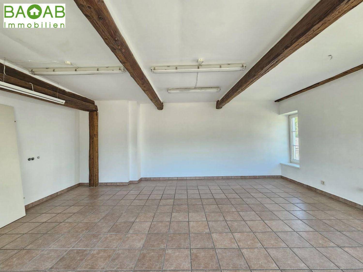 Leerer Büroraum mit Fliesenboden, weißen Wänden, einem Fenster und sichtbaren Holzdeckenbalken, mit Türzugang.