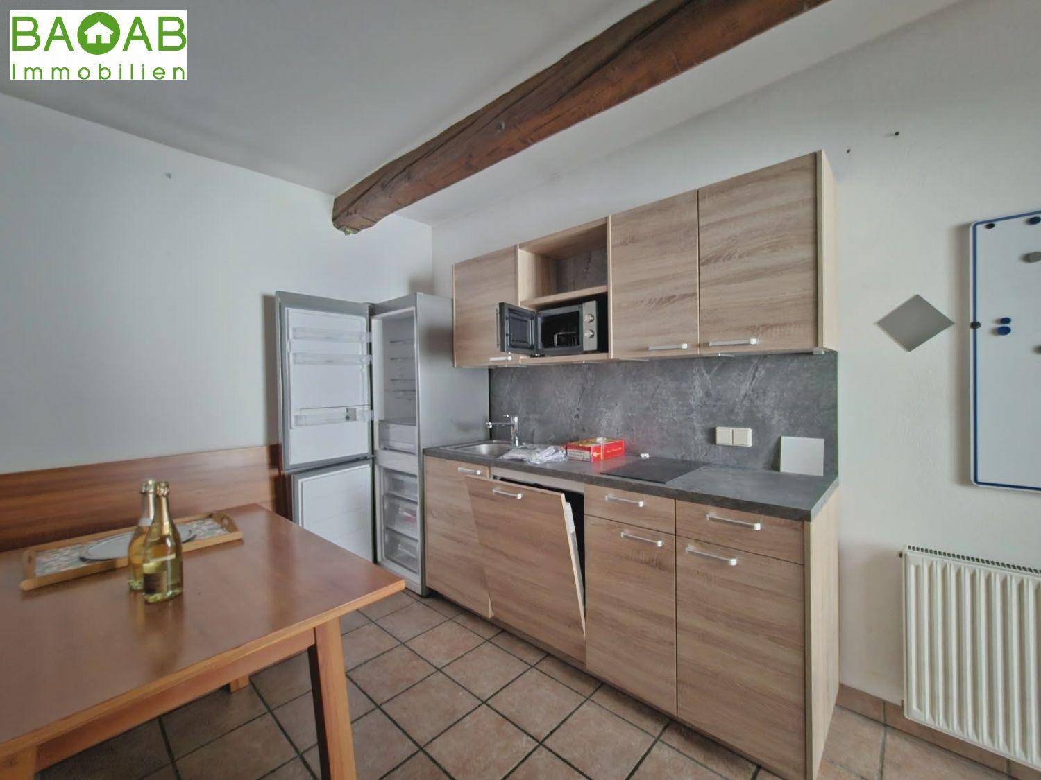 Kleine Einbauküche mit Holzschränken, Induktionskochfeld, Mikrowelle und Kühlschrank, ergänzt durch einen Esstisch.