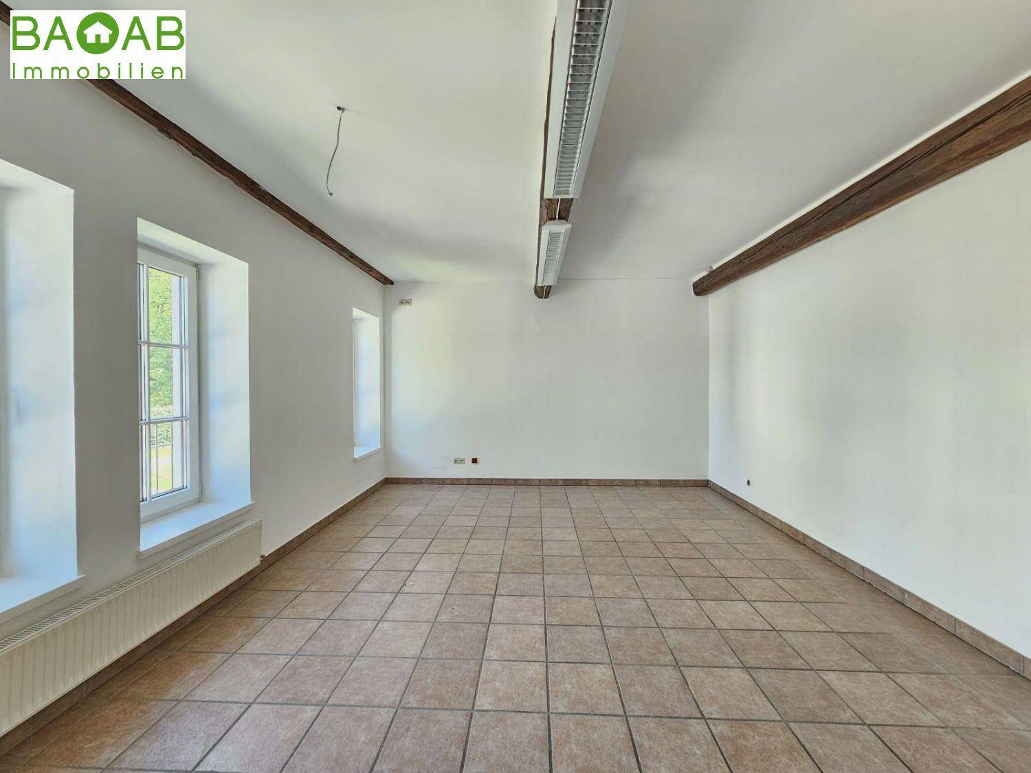Heller, leerer Raum mit Fliesenboden, weißen Wänden und einem sichtbaren Holzdeckenbalken, ideal für Büronutzung.