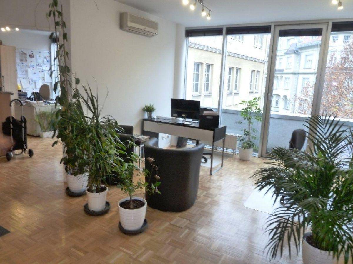 Offener Bürobereich mit mehreren Arbeitsplätzen, vielen Pflanzen und großen Fenstern mit Blick auf die Stadt.