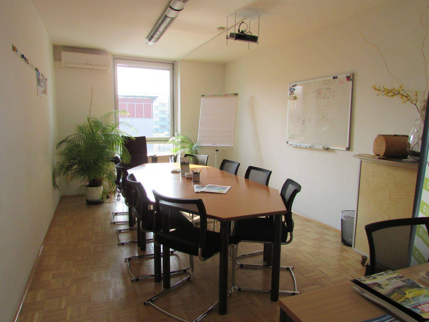 Heller Konferenzraum mit ovalem Tisch, Stühlen, Whiteboard, Fenster und einem Deckenprojektor für Präsentationen.