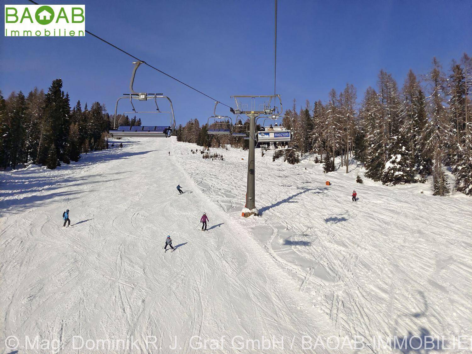 Sonnige Skipiste mit mehreren Skifahrern und einem Sessellift, umgeben von verschneiten Bäumen.
