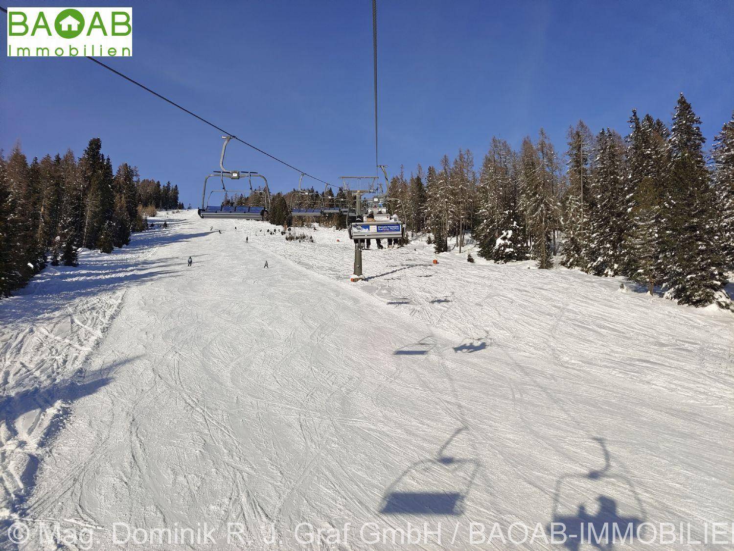 Skifahrer auf einer breiten Piste mit einem Sessellift und verschneiten Bäumen im Hintergrund.