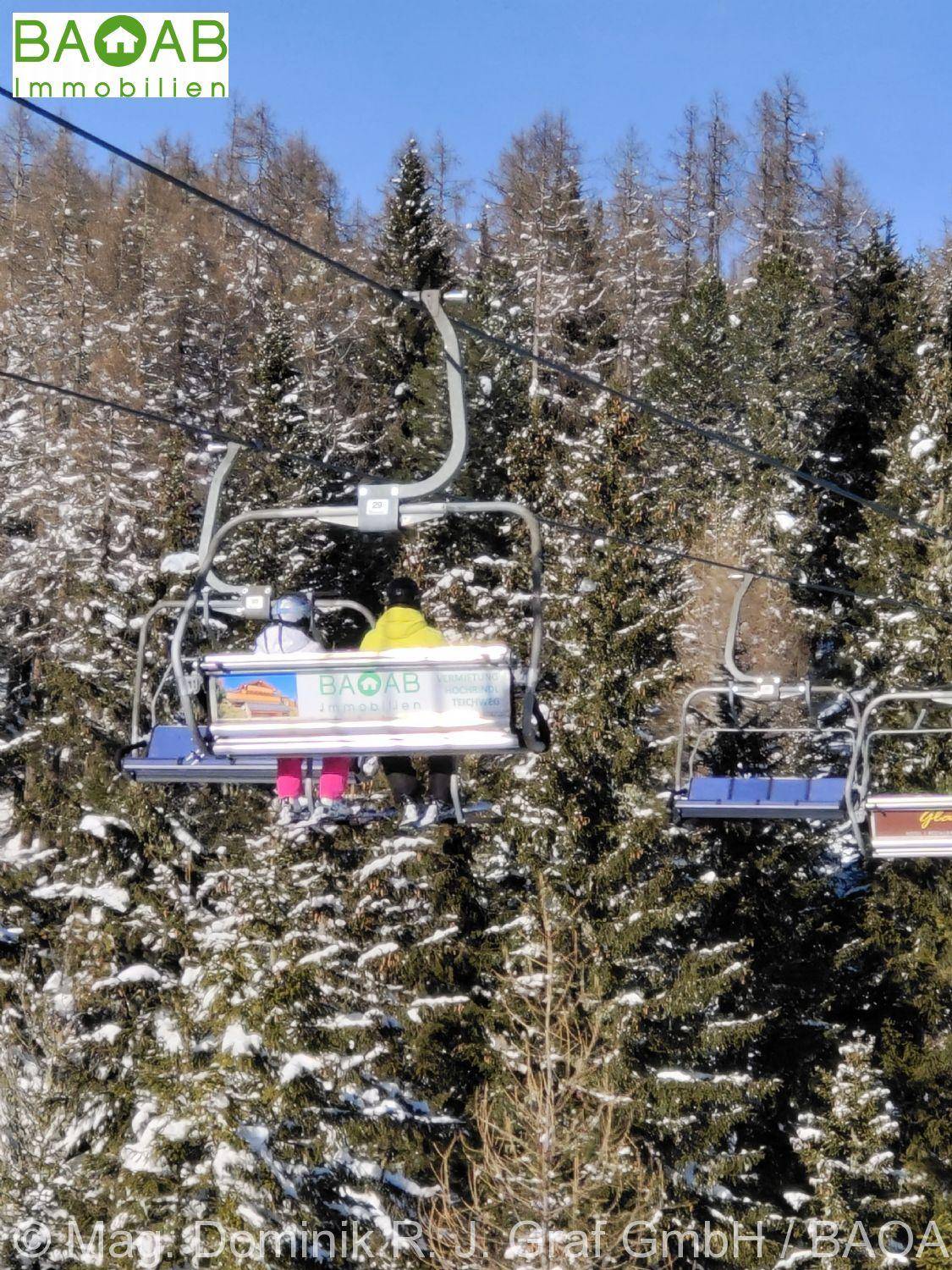 Personen fahren mit dem Sessellift über verschneite Tannenwälder in einem Skigebiet.