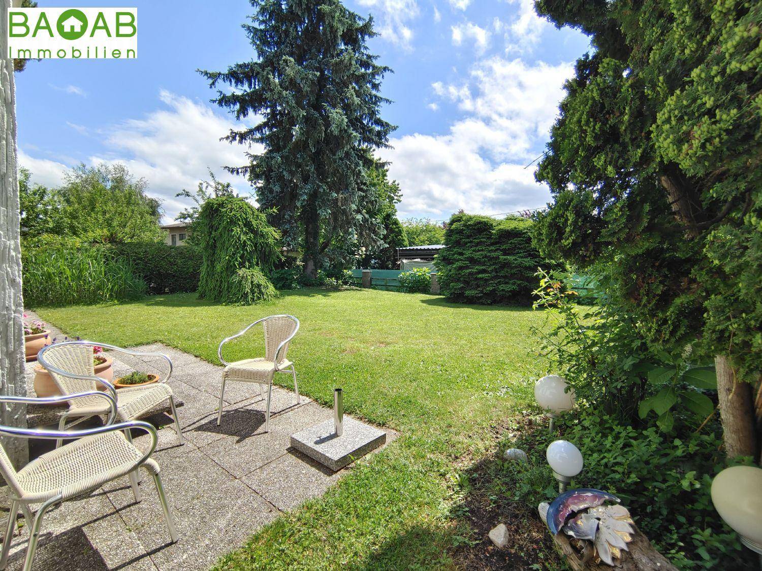 Gepflegter Garten mit Rasenfläche, Sitzgelegenheiten und hohen Bäumen unter blauem Himmel.