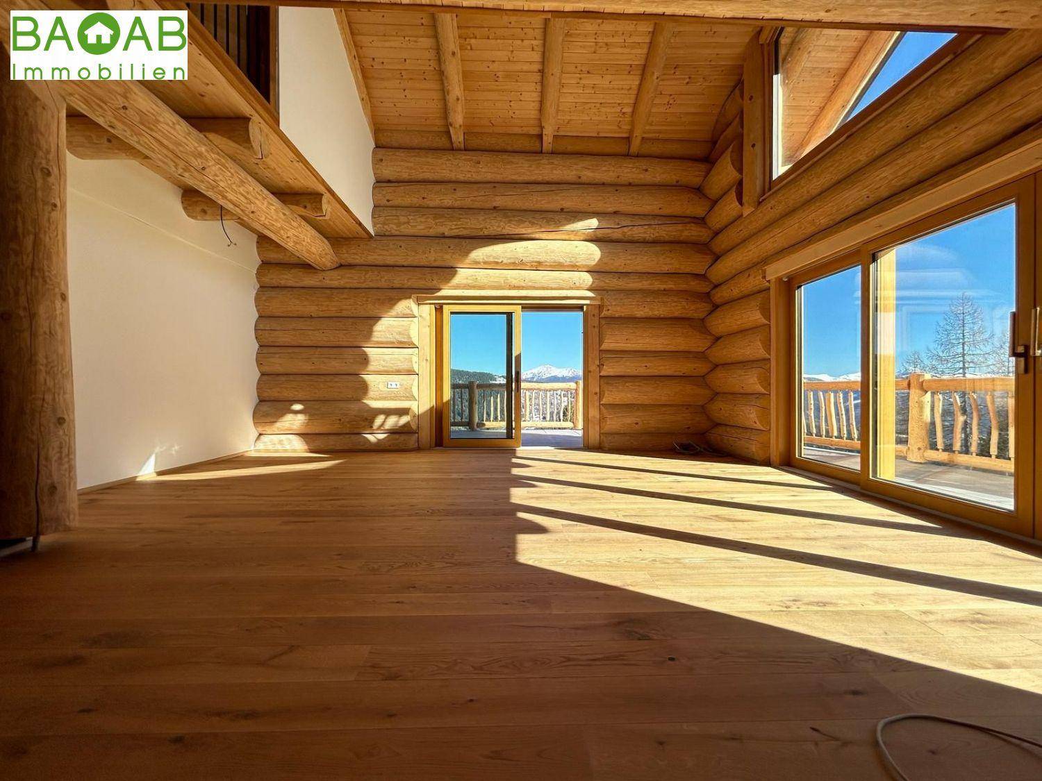 Heller Innenraum eines Holzchalets mit Holzbalken, großen Fenstern und Zugang zu einer Terrasse mit Bergblick.