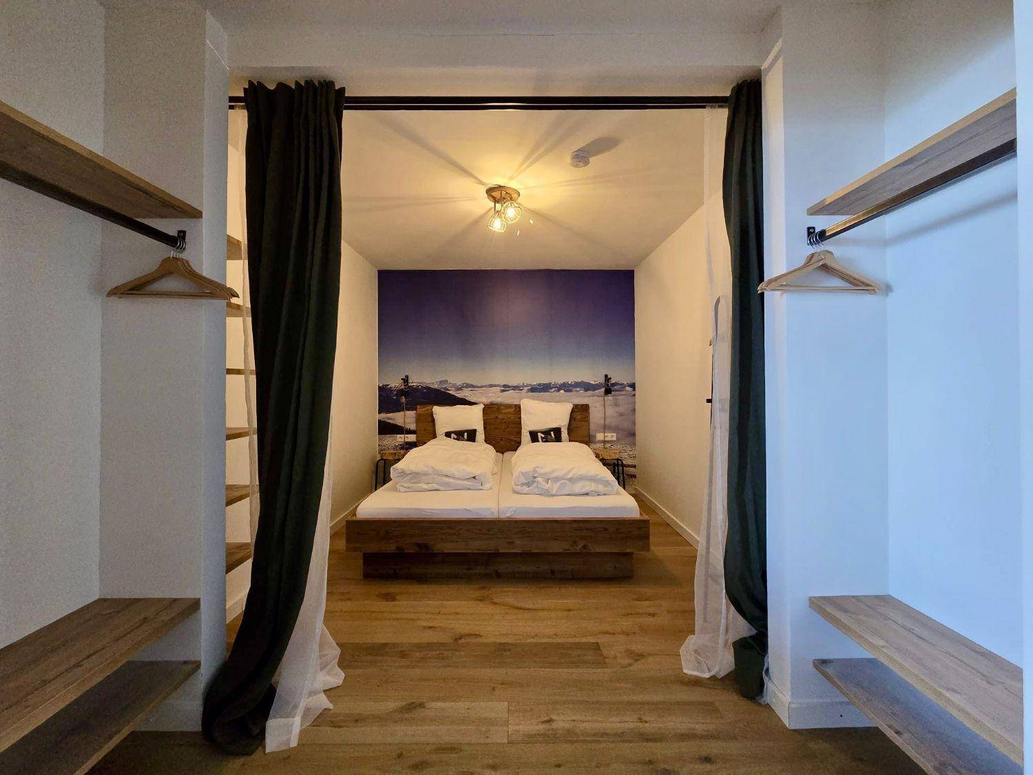 Schlafzimmer mit Holzbett, Bergpanorama-Wandbild und offenem Kleiderschrankbereich.