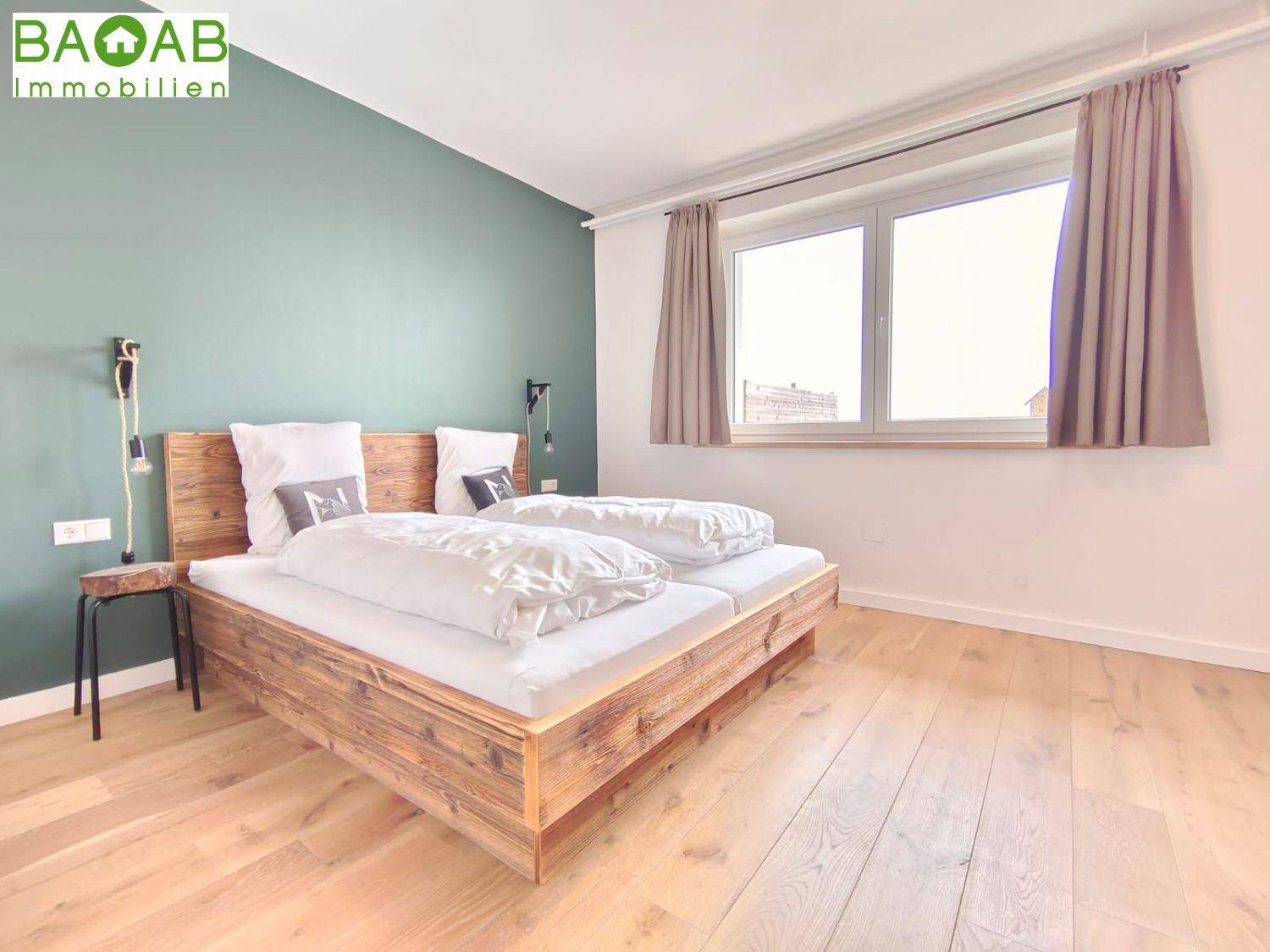 Schlafzimmer mit Holzbett, grüner Akzentwand und viel natürlichem Lichteinfall durch das Fenster.