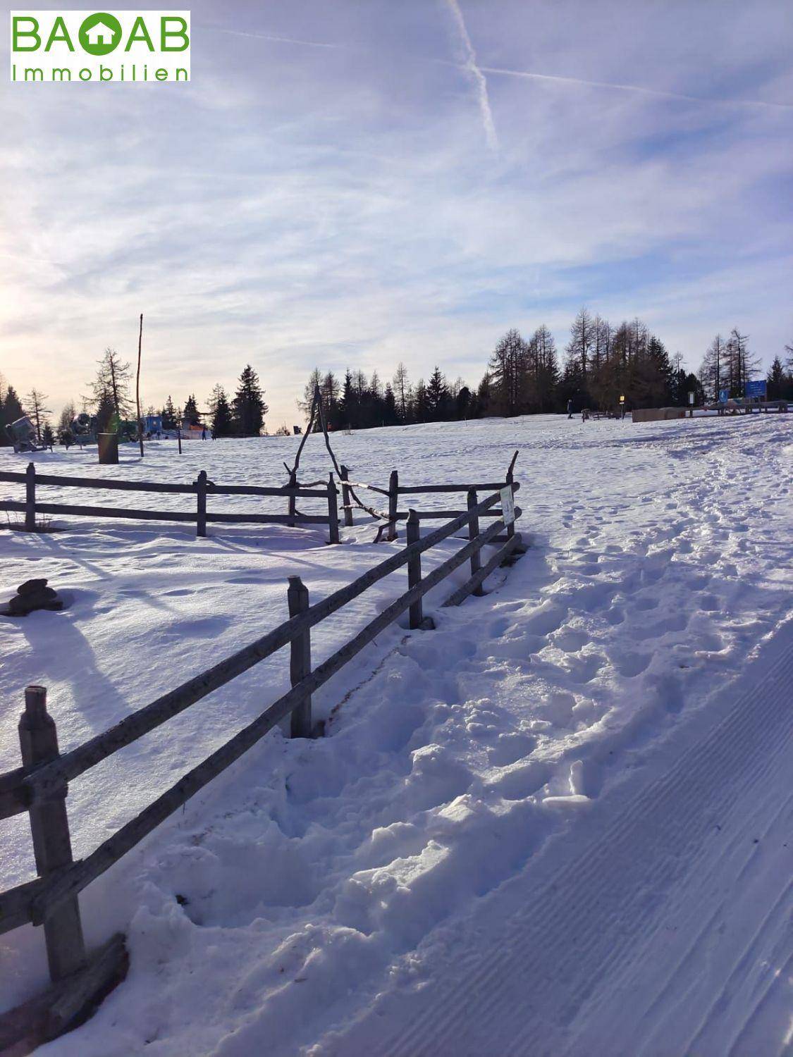Winterliche Landschaft mit verschneiten Feldern, einem Holzzaun und Bäumen unter blauem Himmel.