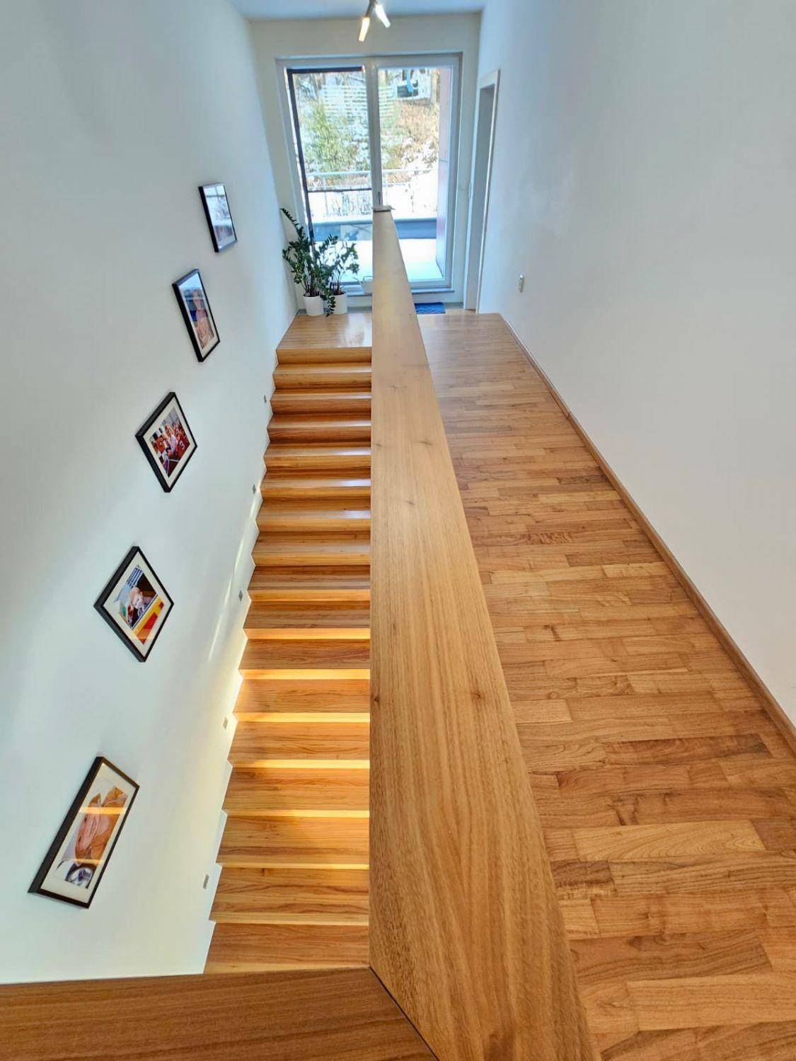 Helles Treppenhaus mit moderner Holztreppe, Glasgeländer und Holzboden.