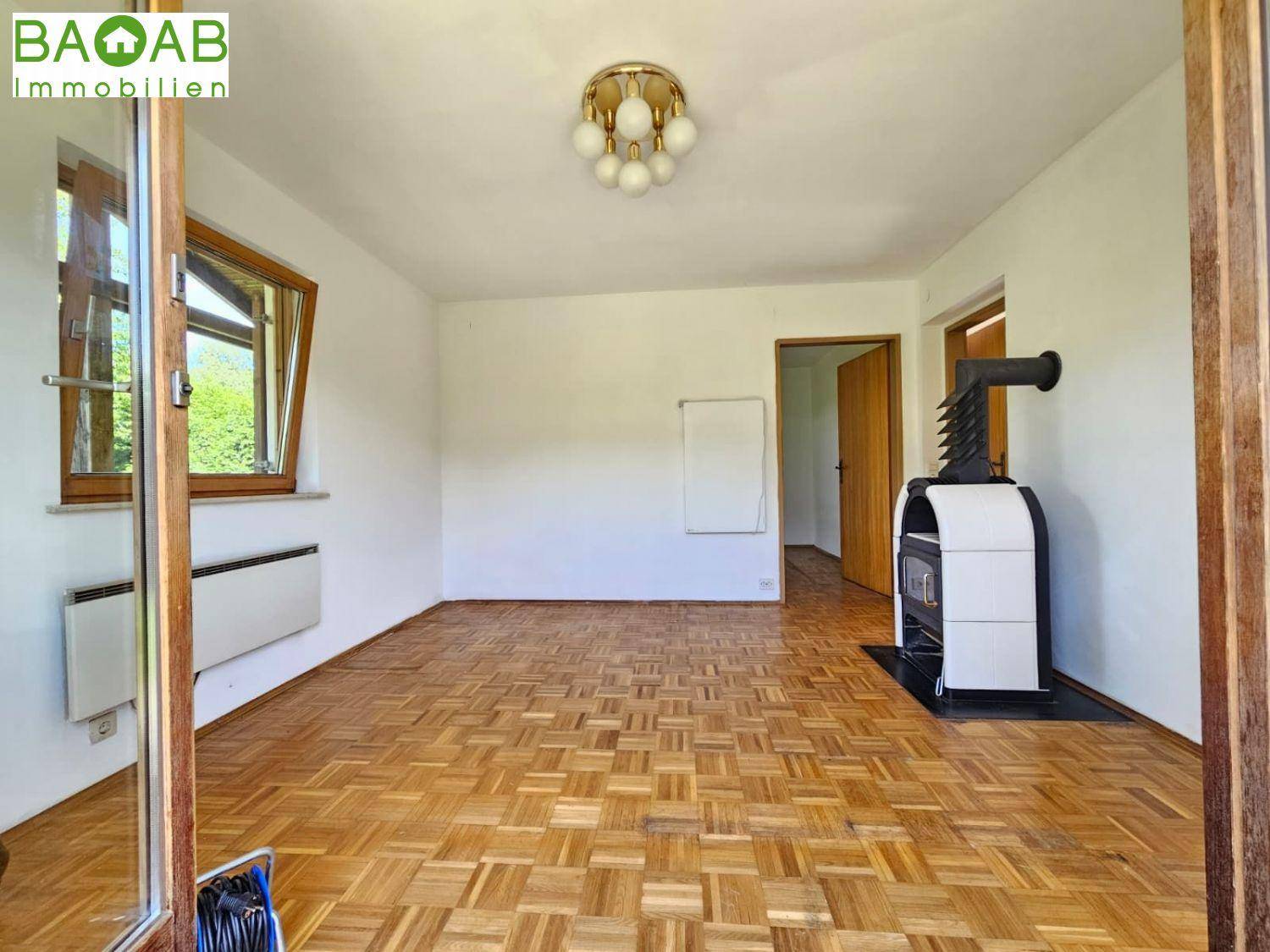 Heller Raum mit Parkettboden, Kaminofen und einem Fenster mit Blick auf die Natur.