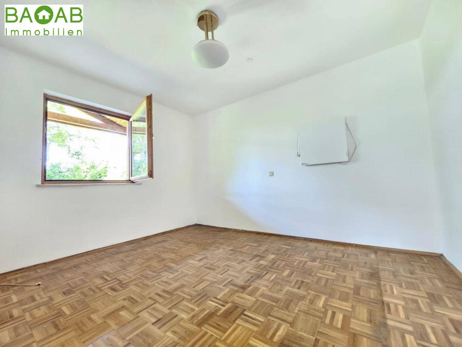 Leeres Zimmer mit Parkettboden, weißen Wänden und einem Fenster mit Blick ins Grüne.