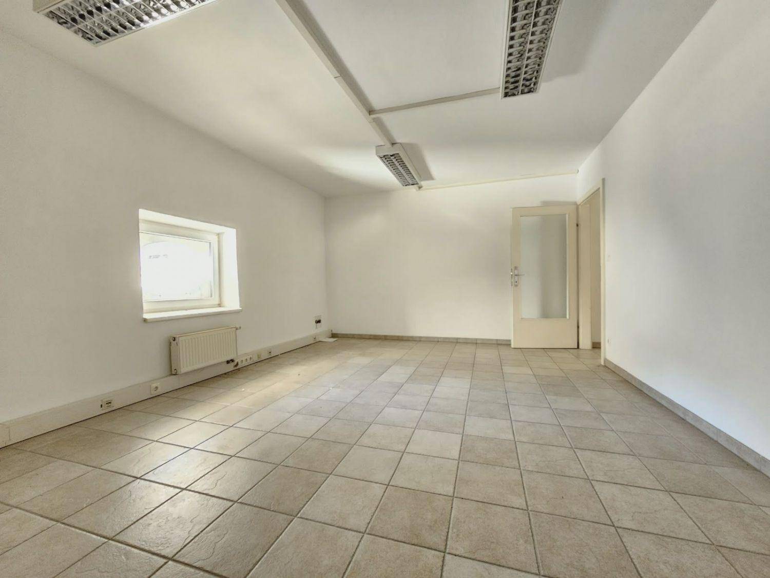 Heller, leerer Büroraum mit Fliesenboden, einem Fenster und funktionaler Deckenbeleuchtung.