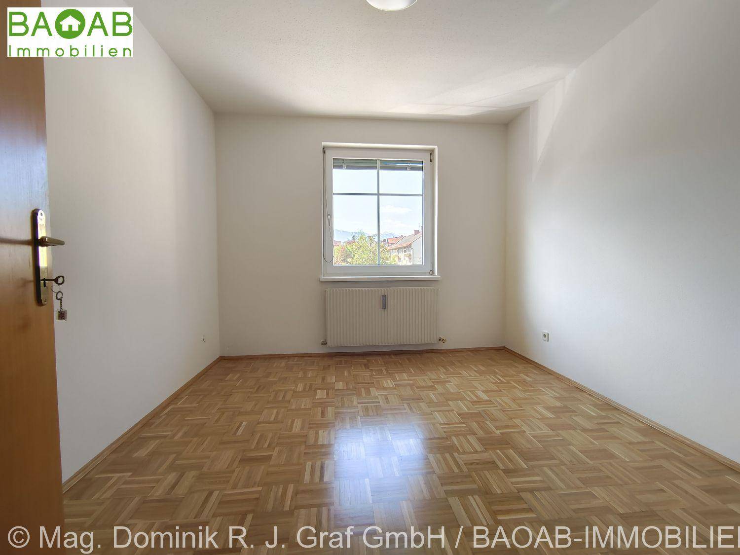 Leeres Zimmer mit Parkettboden, weißen Wänden und einem Fenster mit Heizkörper darunter.
