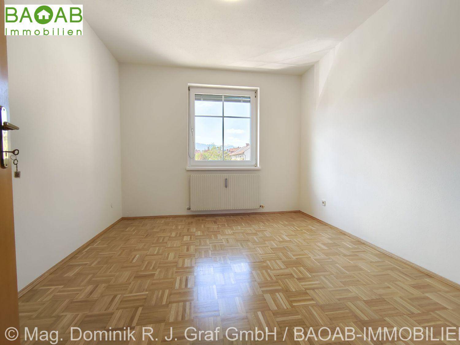 Leeres Zimmer mit Parkettboden, weißen Wänden und einem Fenster mit Heizkörper darunter.