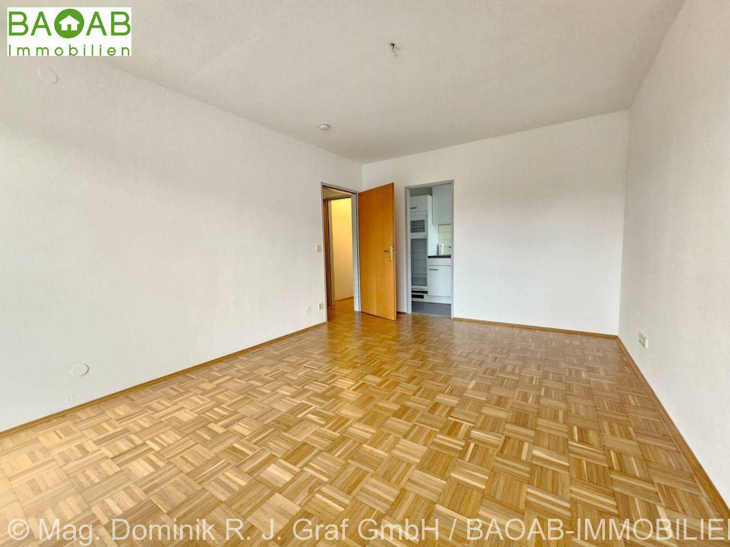 Heller, leerer Raum mit Fischgrätparkettboden und Zugang zu einem weiteren Zimmer sowie einer Küche.