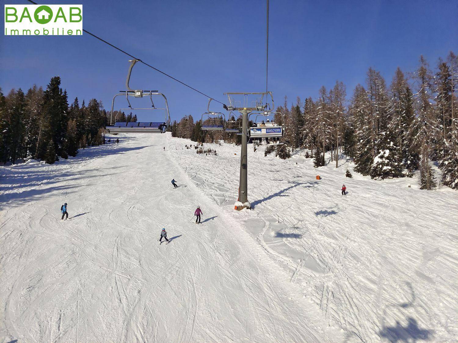 Weite Skipiste mit mehreren Skifahrern und einem Sessellift, umgeben von winterlichen Wäldern.